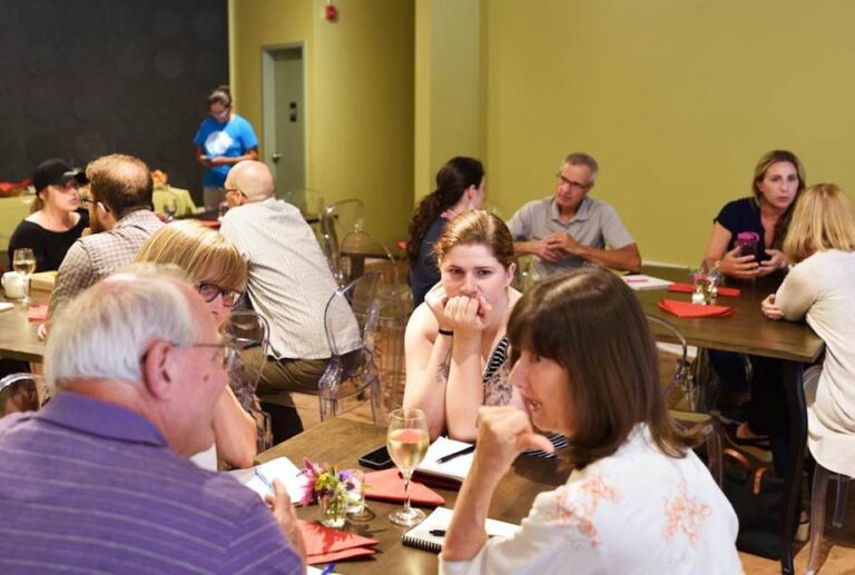 A room of people sit at tables discussing ideas during a summer institute event at The Working Centre's Fresh Ground location
